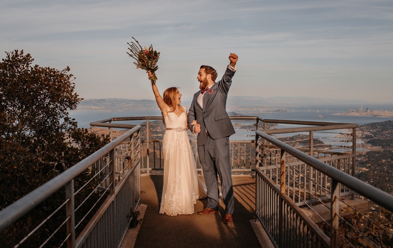 Elopement photographer, bride and groom throw hands in the air, at an overlook of the city