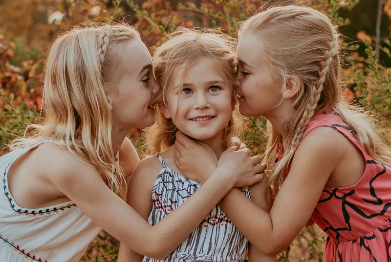 Family Photographer, two sisters lean in to kiss their younger sister, the younger sister is amused