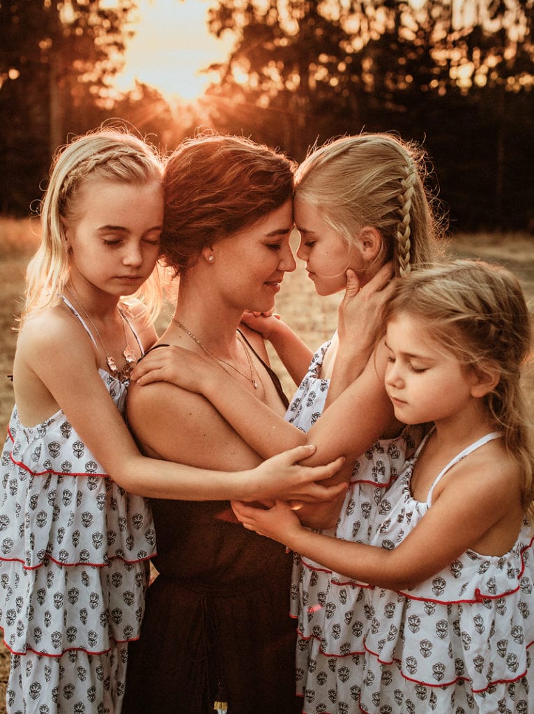 a mother stands and leans into her three daughters outdoors at golden hour