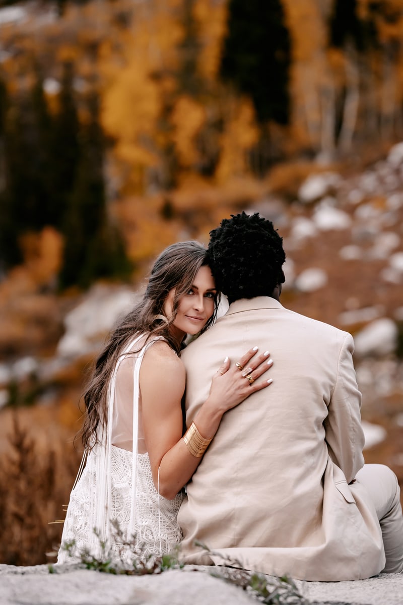 Elopement photographer, a woman leans on her husbands shoulder happily as they sit on a rock's edge in the mountain forests
