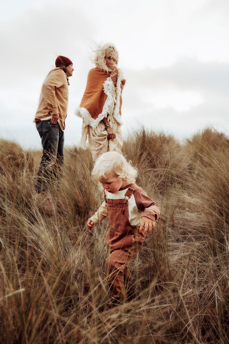Family Photographer, mom and dad watch over their toddler son as he wanders through beach seagrass