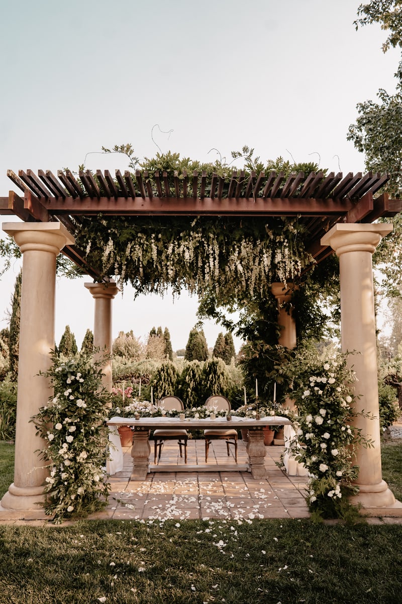 wedding photographer, a table is setup outdoors adorned with flowers beneath a pergola for a reception