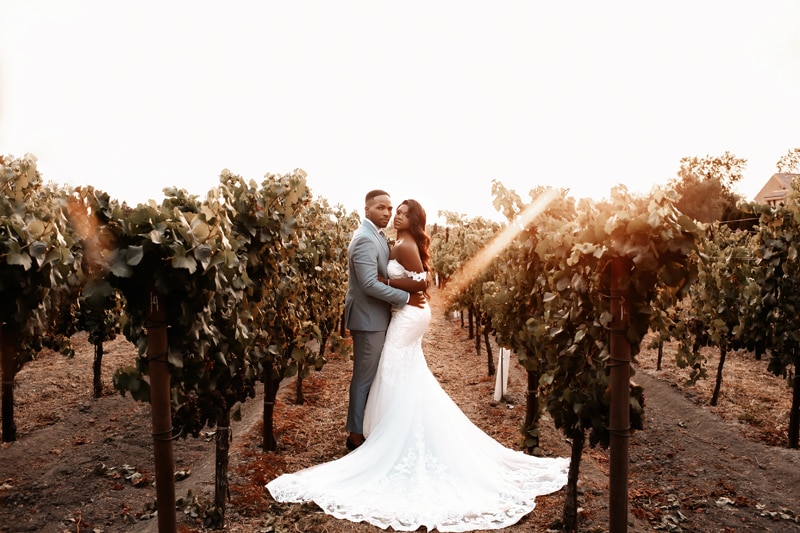 Wedding photographer, husband and wife in their tux and wedding dress, walk through a vineyard at golden hour