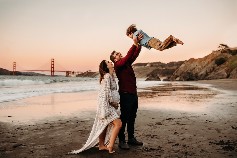 Maternity Photographer, an expecting mother stands on the beach with her husband, he is a dad and swing his son in the air. The golden gate bridge is behind them