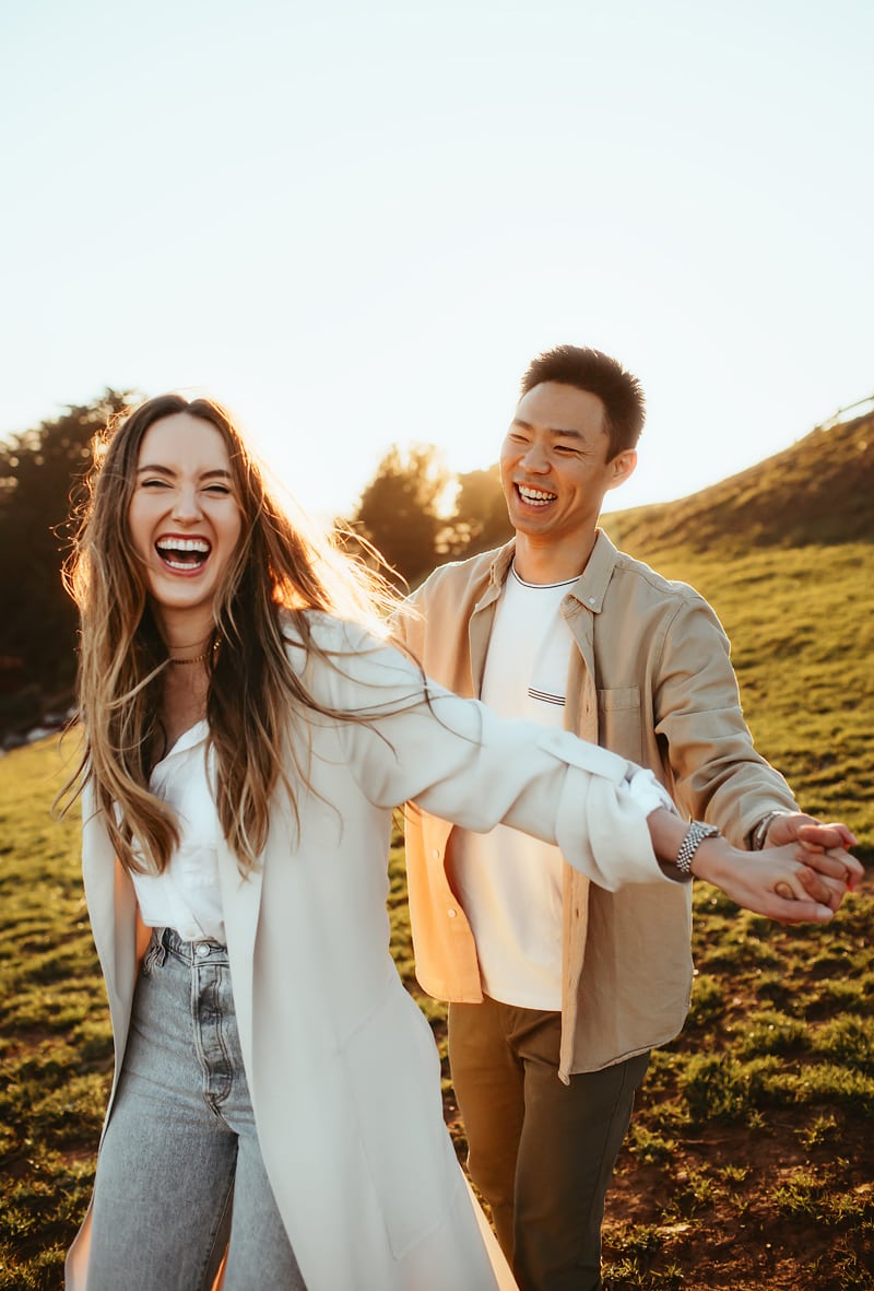 Couples Photographer, man and woman hold hands laughing as they walk through grassy hillside