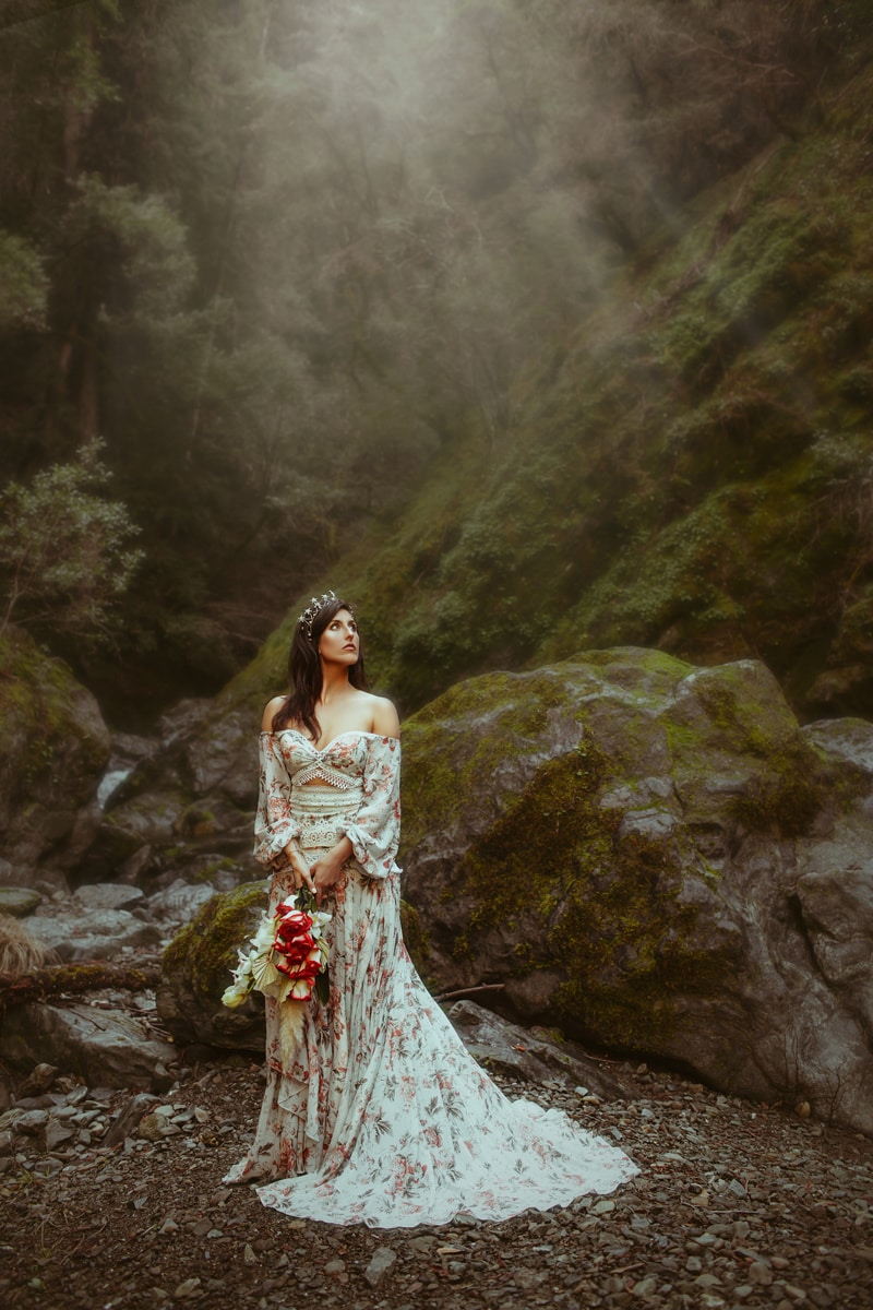 Wedding photographer, a bride stands among large mossy rocks, she wears a dress with floral patterns