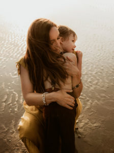 A mother hugs her son, they stand in the sand at the beach near the water