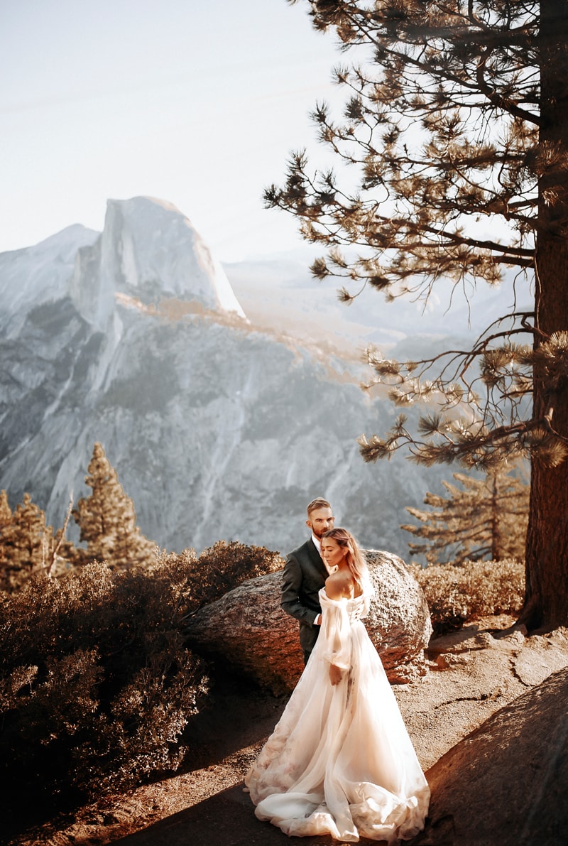 wedding photographer, a bride and groom stand on a mountain trail, Yosemite's half dome is behind them