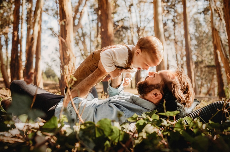 Family Photographer, a young father admires his son in his arms as he lays in a grove of trees