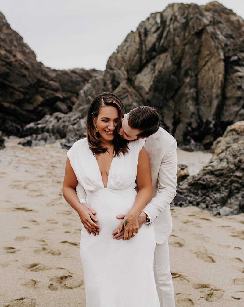 Elopement photographer, a husband hugs his new wife from behind as they stand on a sandy beach