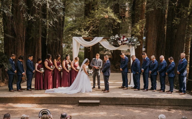wedding photographer, a man and woman bow their heads during their wedding in the forest, all the bridesmaids and groomsmen are lined up beside them