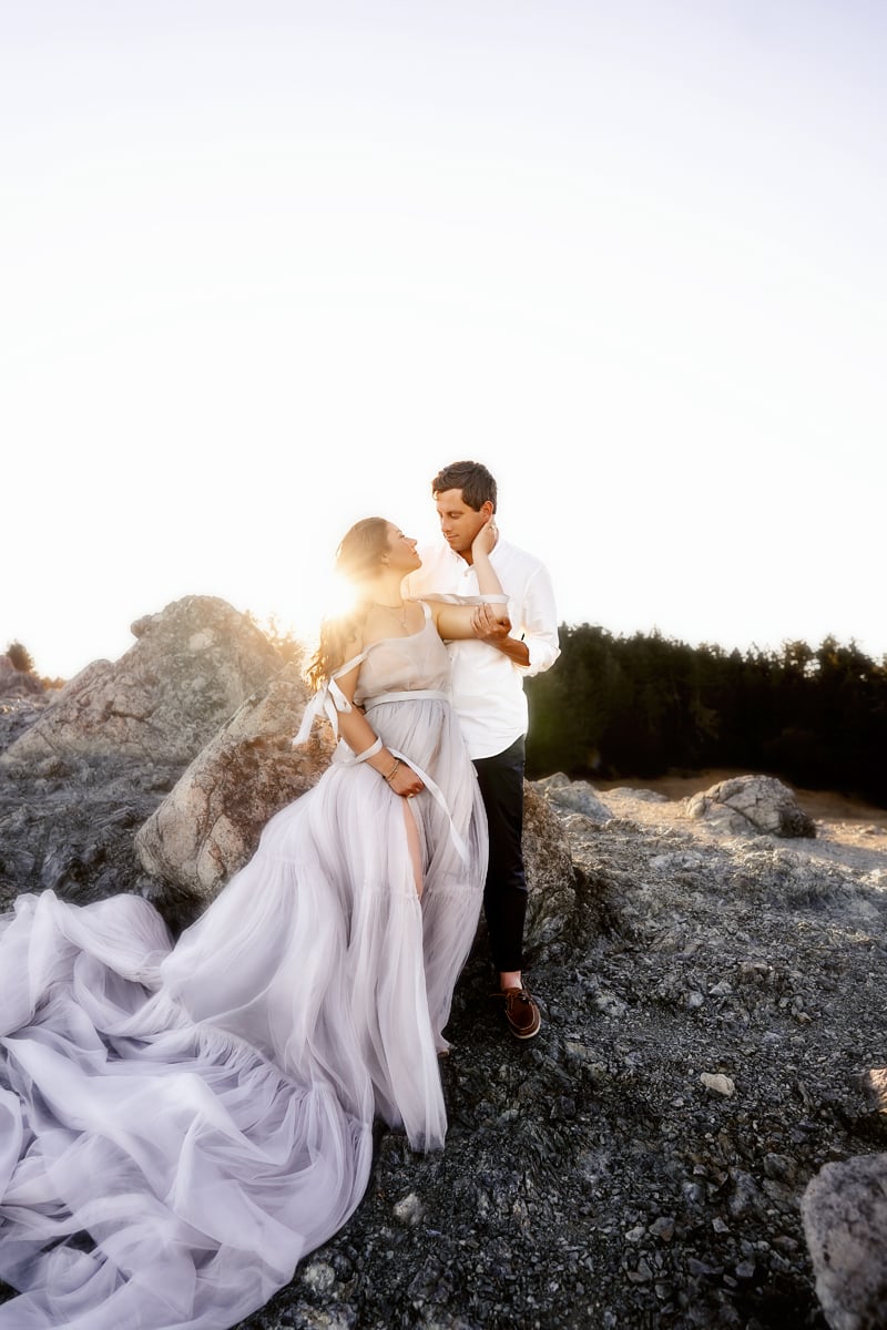 Couples Photographer, man holds woman, she wears long flowing dress, they are among boulders outdoors