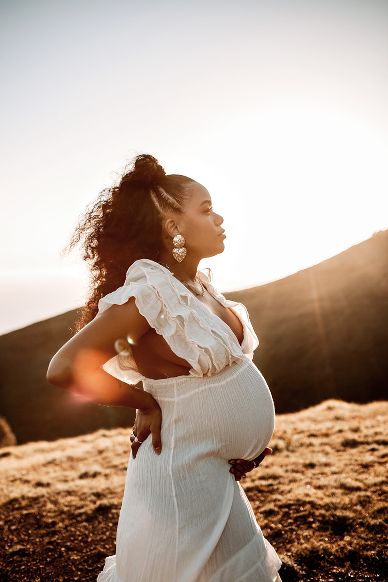 Maternity Photographer, an expecting mother stands confidently on a hillside, the sun shines brightly behind her