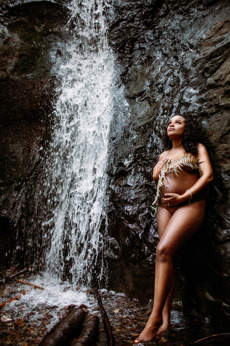 Maternity Photographer, an expecting mother holds her belly as she stands in the midst of a waterfall, she is only covered by a palm branch