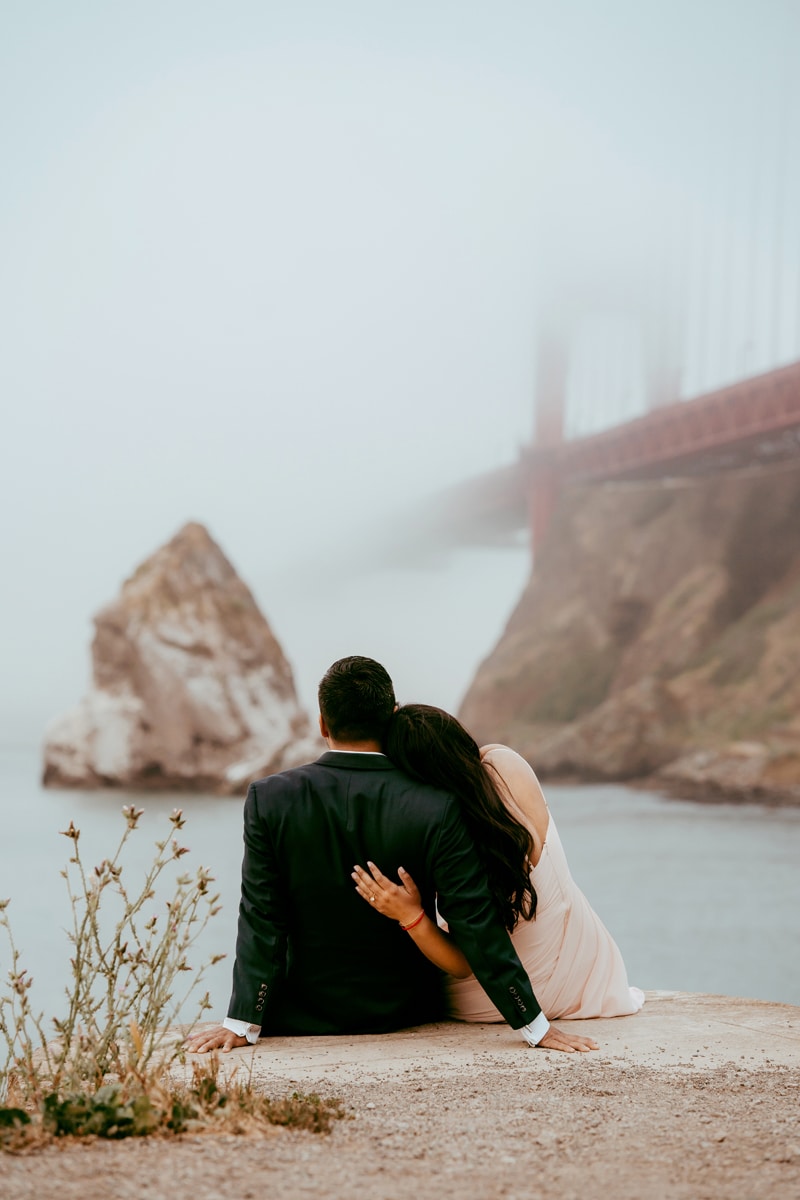 Couples Photographer, a man and woman sit near the Bay, The Golden gate bridge before them in the fog