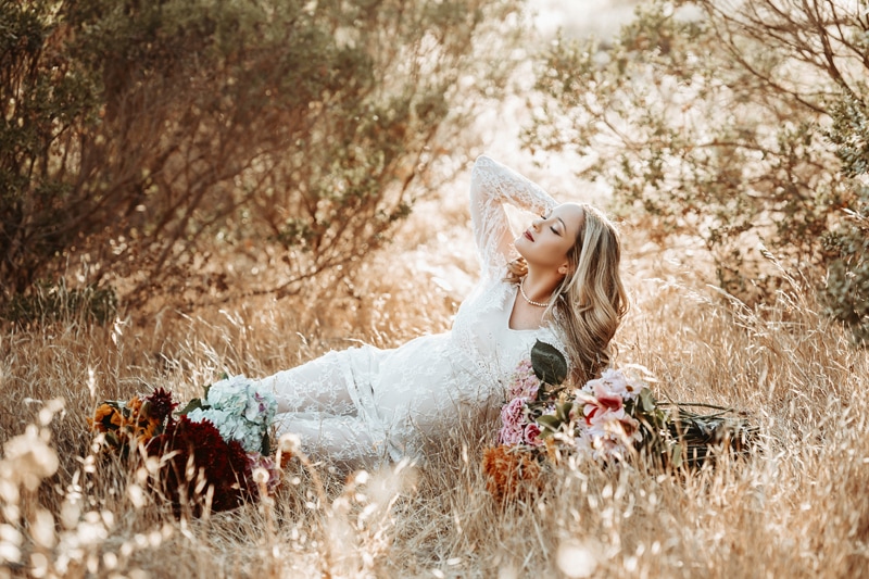 Maternity Photographer, a woman expecting child lays in dry grass with a bouquet of flowers beside her