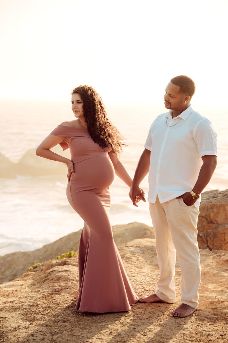 Maternity Photographer, husband admires expecting wife as they hold hands near the ocean