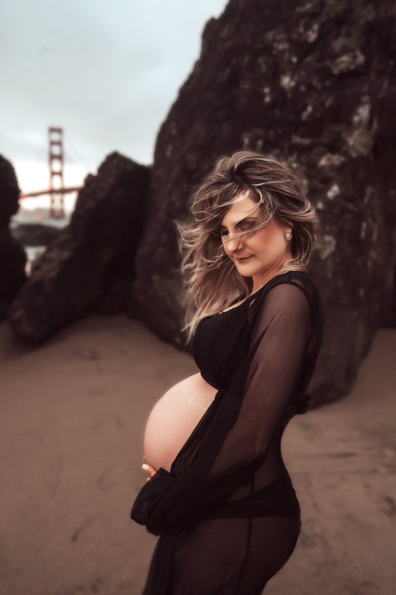 Maternity Photographer, an expecting mother stands on the beach, the golden gate bride in the distance behind her