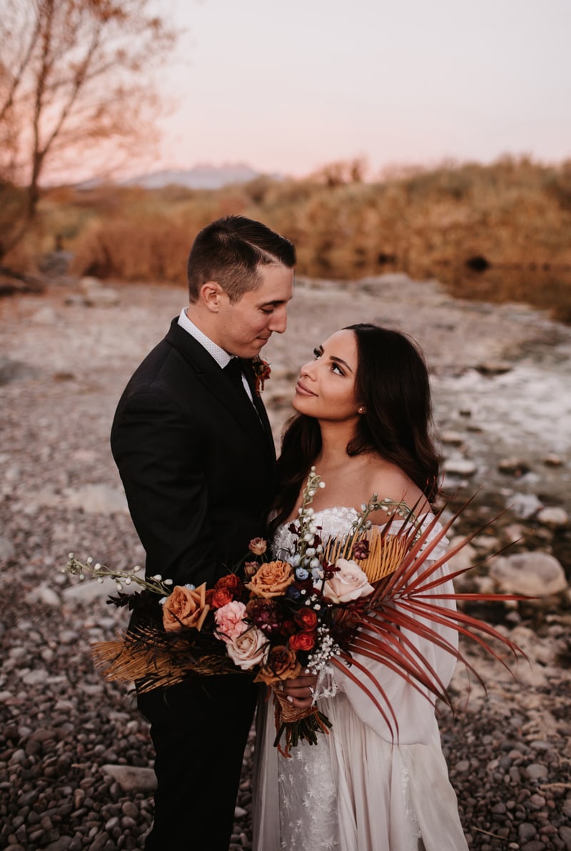 wedding photographer, a bride and groom gaze into each other's eyes, she holds a bouquet near the river's edge