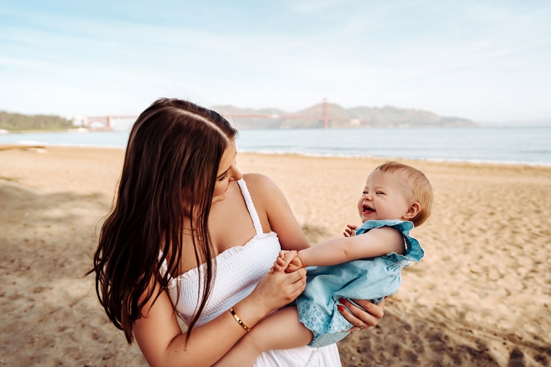Family Photographer, a young mother plays with laughing baby at the beach, the golden gate bridge behind them