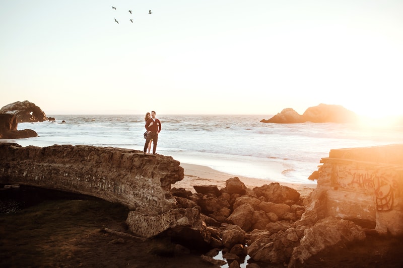 Couples Photographer, woman hugs man from behind as they stand on rock wall near ocean