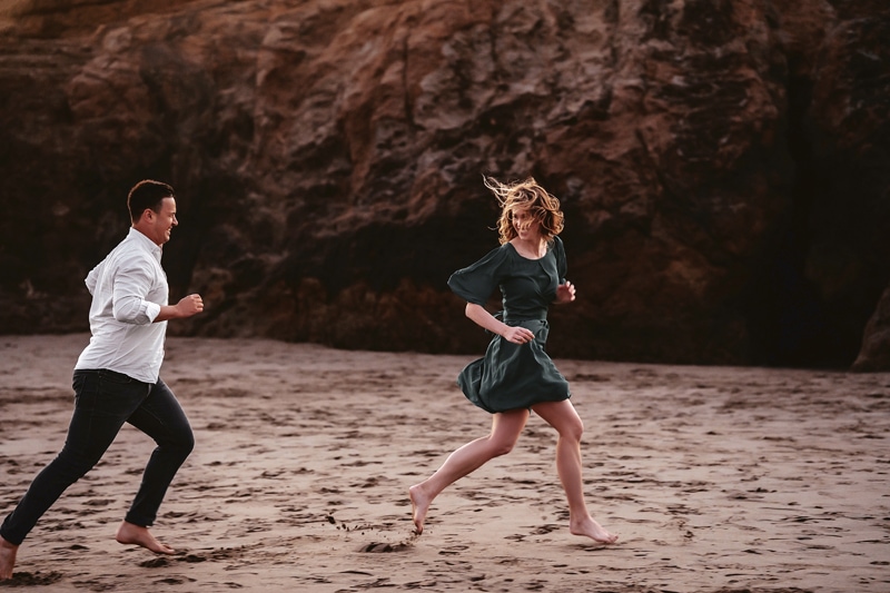 Couples Photographer, man chases woman as she smiles on the beach
