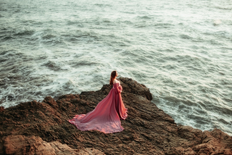 Maternity Photographer, a woman in long flowing dress is expecting and stands on the rocks near the ocean