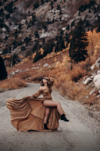 a woman stands on a mountain road, she holds her dress as it flows in the wind